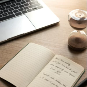 A notebook, laptop, and a hourglass on a table