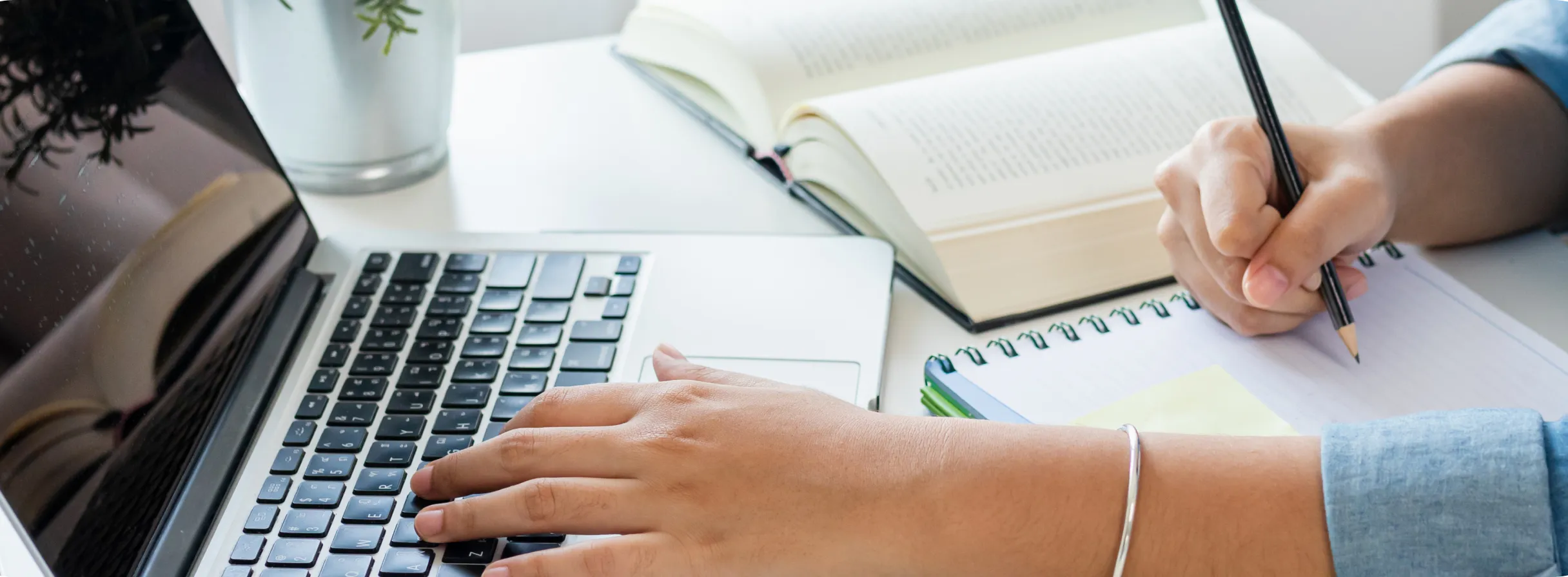 A person typing on a laptop while writing on a notebook