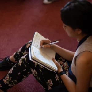 A woman sitting on the floor writing on her notebook.