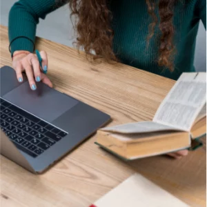 A person holding a book while doing something in her laptop