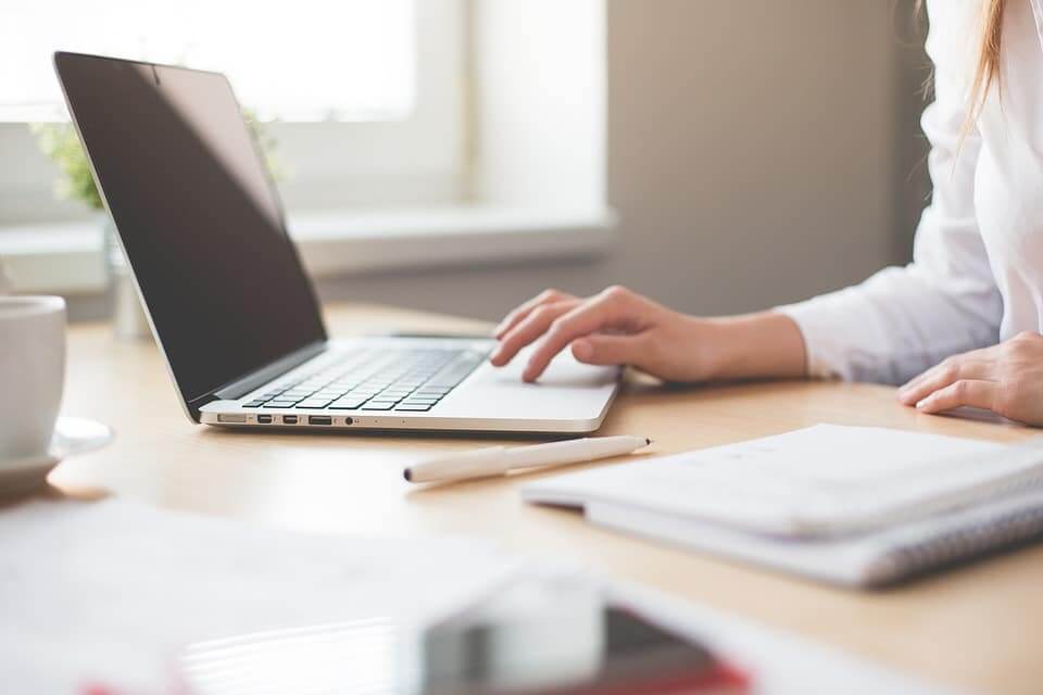 A person hovering on her laptop with a notebook and a pencil on her table.