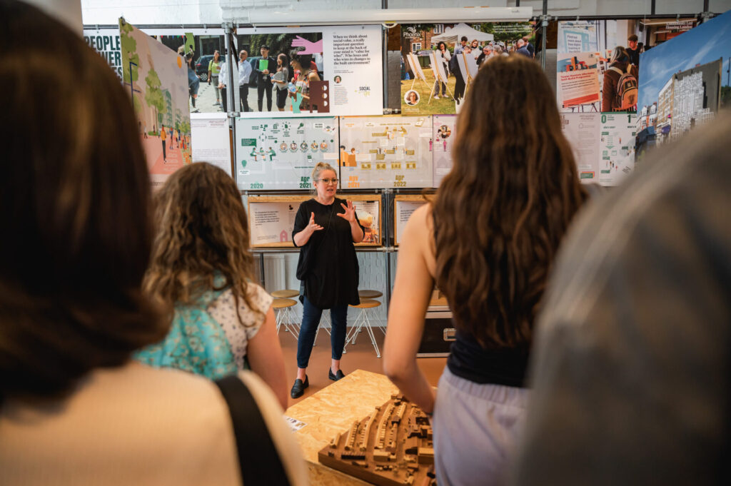 In the foreground, the blurred back of several year 12 work experience students, looking on as a woman in front of a whiteboard explains architecture concepts