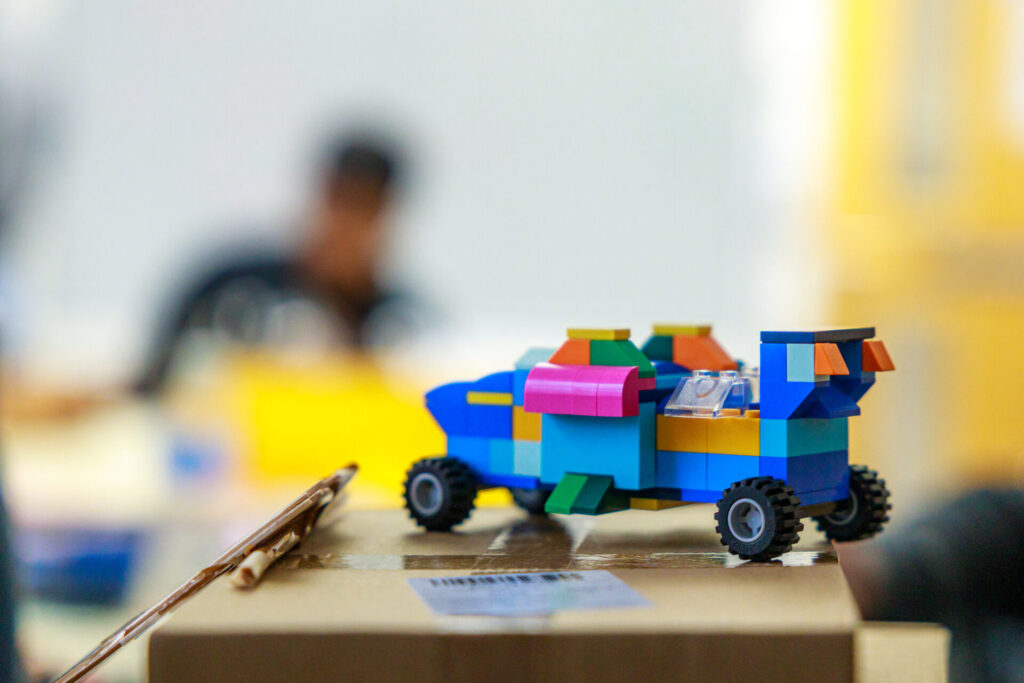 A colourful, lego-built and engine powered toy car in the foreground, with a year 12 student on an engineering work experience programme blurred out in the background