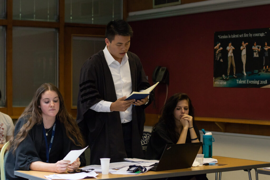 As part of law work experience, a year 12 student stands up in a shirt and gown in a courtroom to read aloud. He is surrounded by two classmates who listen on.