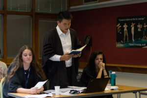 As part of law work experience, a year 12 student stands up in a shirt and gown in a courtroom to read aloud. He is surrounded by two classmates who listen on.
