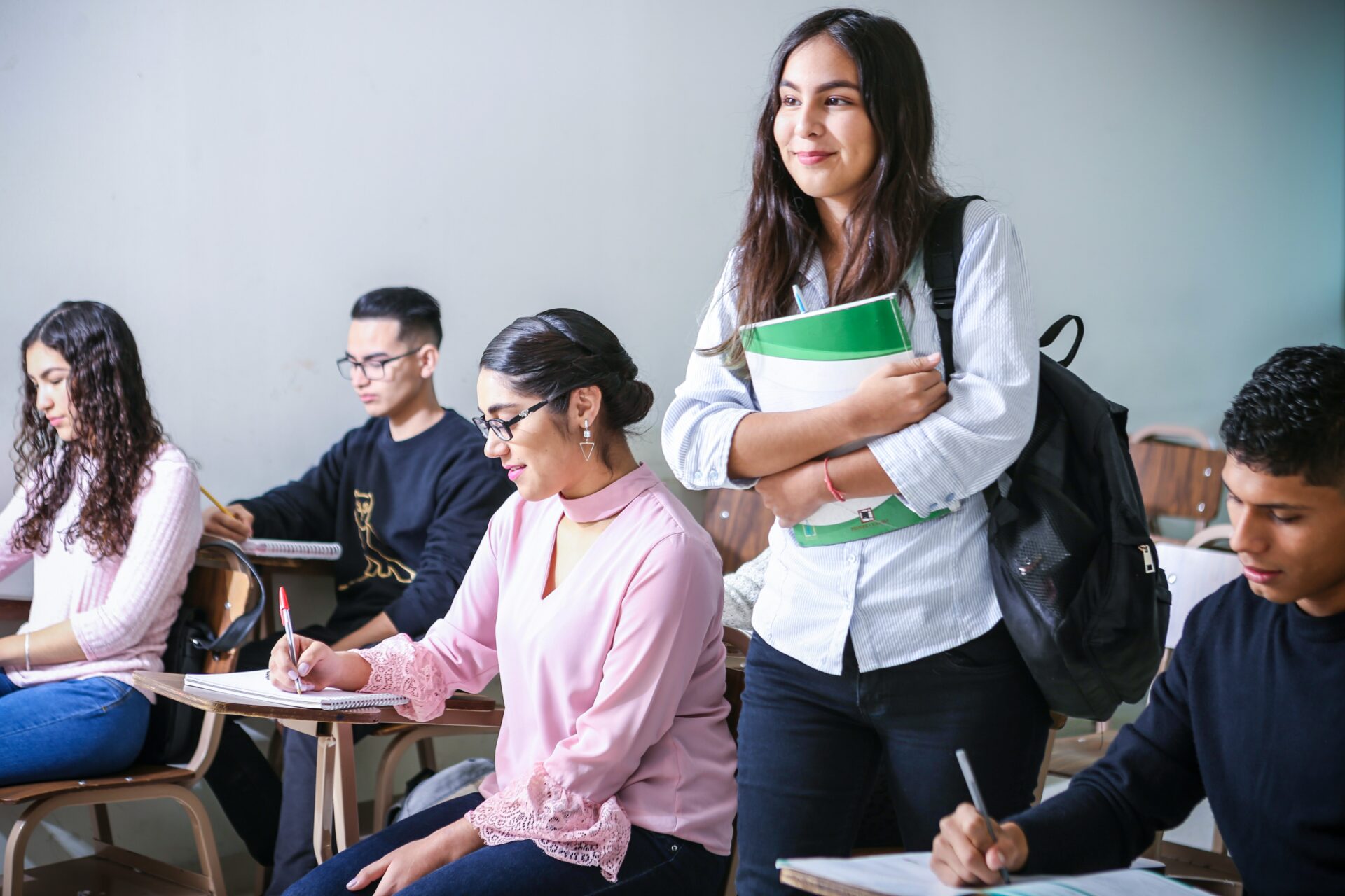 Young, smiling students participate in prestigious summer programs, with a female student standing holding books, while others are seated and taking notes.