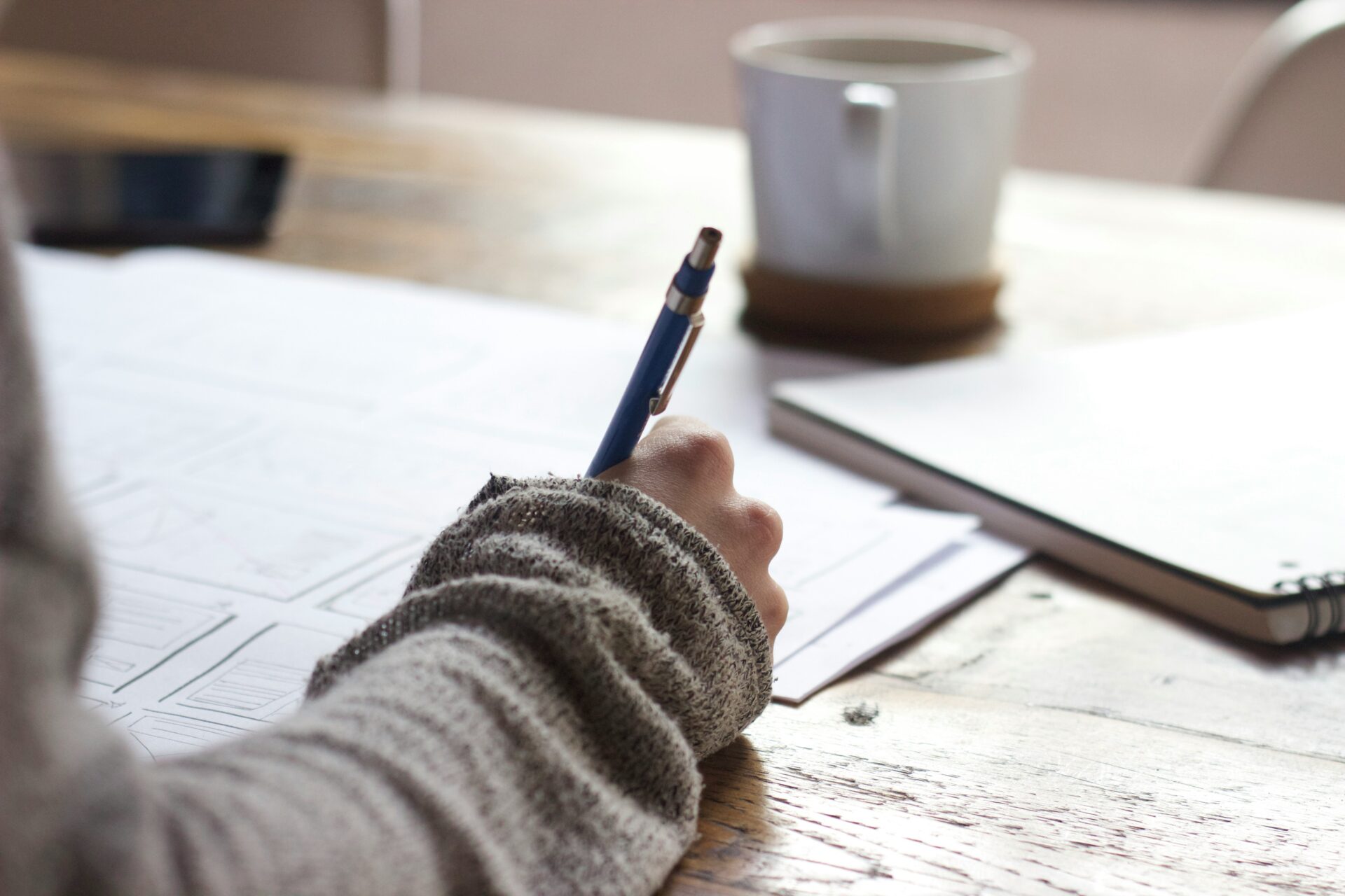 A high school student's hand is shown holding a pen and writing on paper at a wooden desk, with a mug and notebook in the background, illustrating the focus required for the best summer writing programs for high school students.