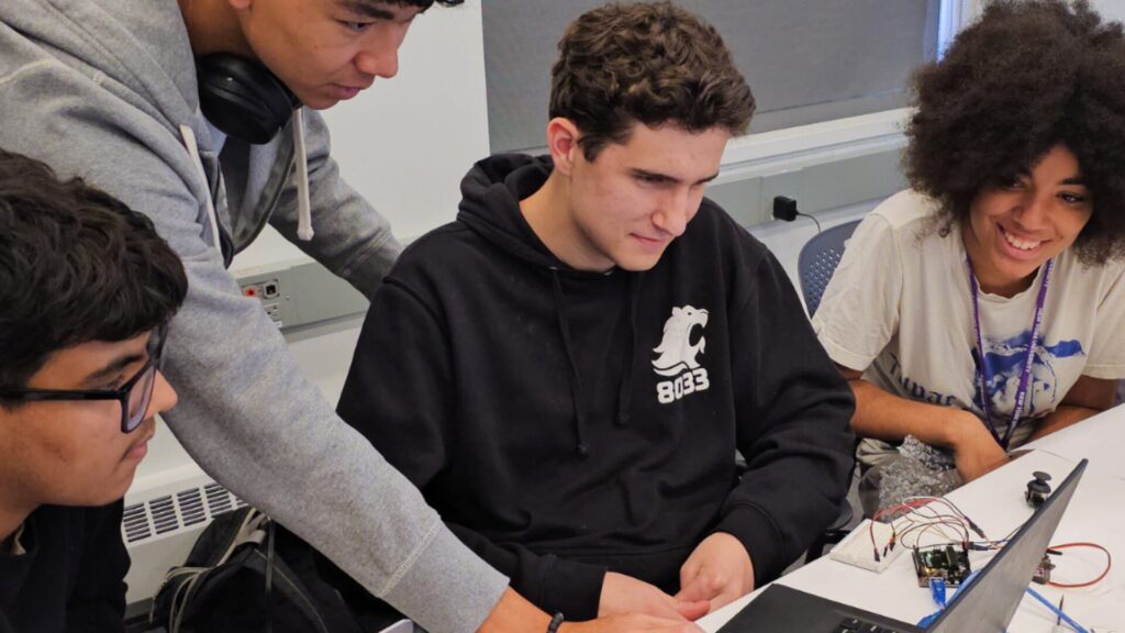 A group of students sitting at the edge of a table and looking at something on a laptop.