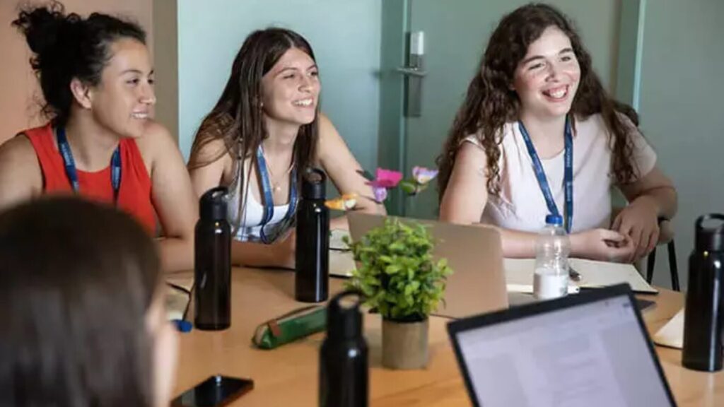 A group of high school students sitting around a table with laptops and water bottles on it, listening to someone speaking.