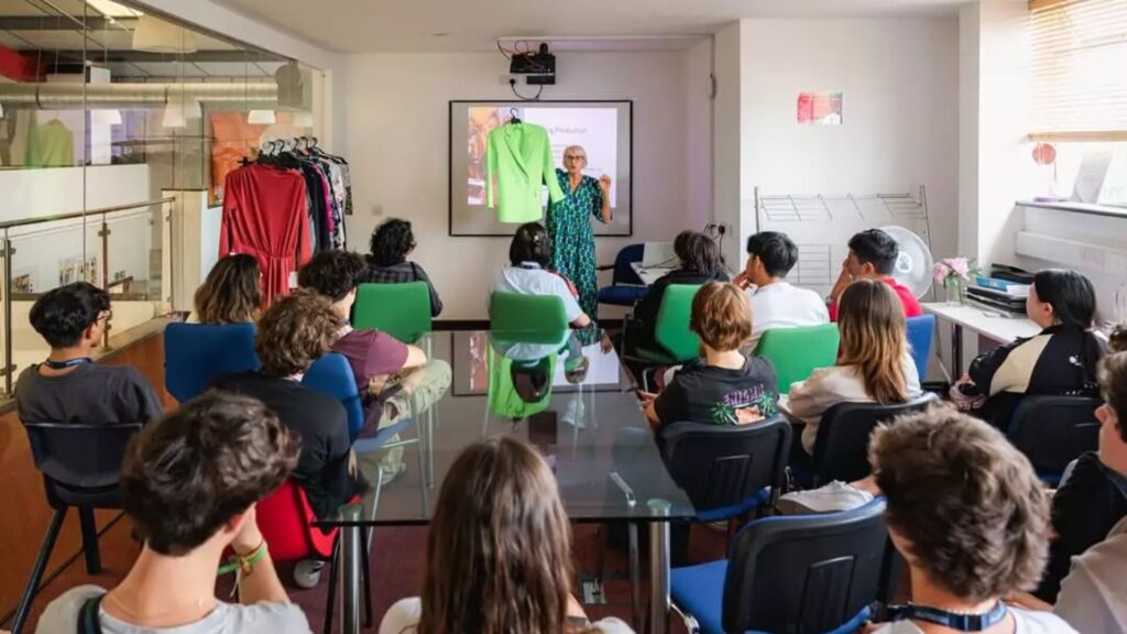 A fashion instructor hosting a class, holding up a green coat as they explain to a room full of seated students.