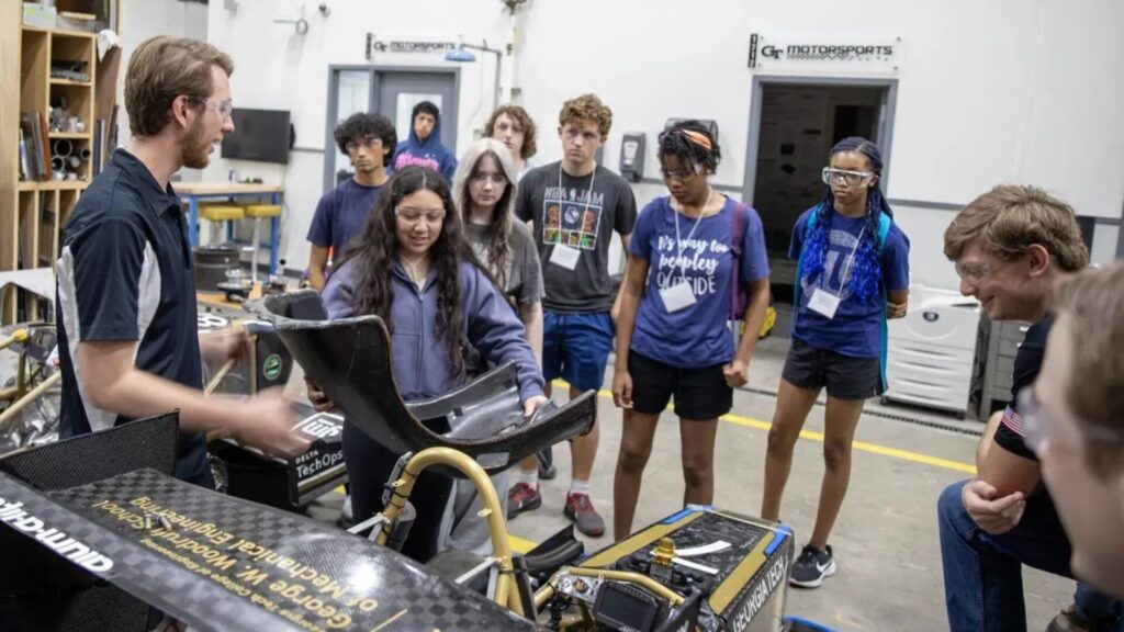 A Georgia Tech engineering student holds up a carbon-fiber race car seat while explaining its design to a group of high school students wearing safety glasses and name badges, gathered around a partially assembled Formula SAE race car inside the Georgia Tech Motorsports workshop.