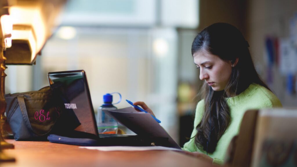 A student attending an online on their laptop and taking notes using a blue pen