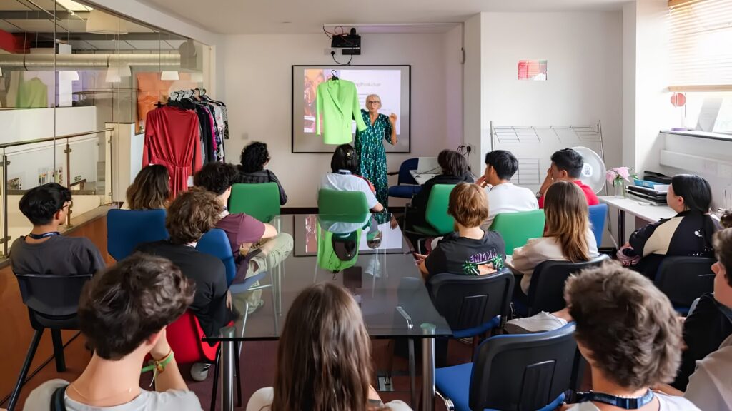 A fashion instructor hosting a class, holding up a green coat as they explain to a room full of seated students.