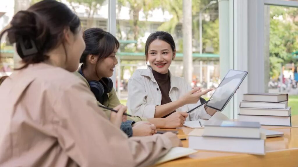 A trio of students sitting around a table lined with books and discussing something.