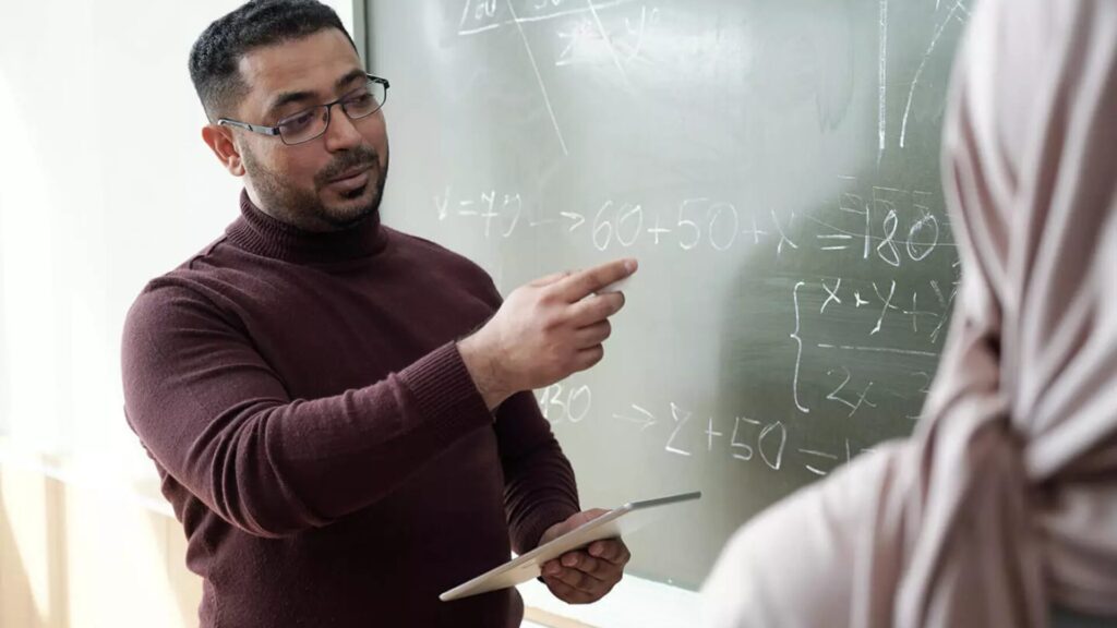 A mathematics instructor gestures at a chalkboard with equations written on it as they hold a tablet and explain something to a high school student.