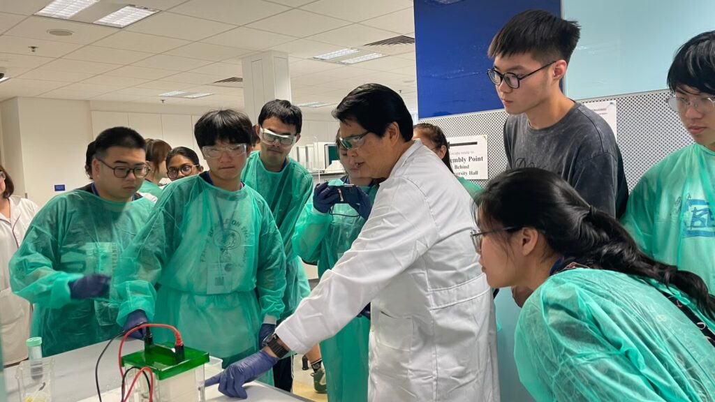 A group of students in lab uniforms look on as a teacher explains a concept to them in a lab.