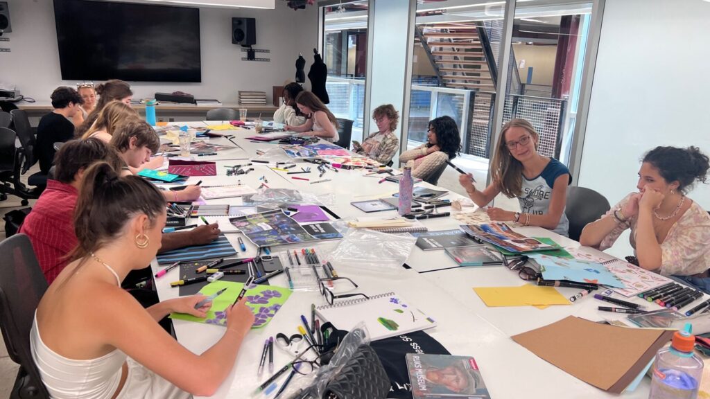 A group of student sitting around a table with notebooks, markers, and pens on the table