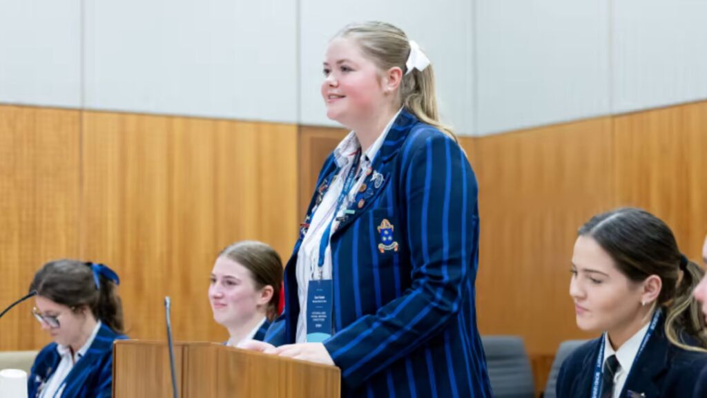 Students wearing blue blazers and sitting, with one student standing behind a podium