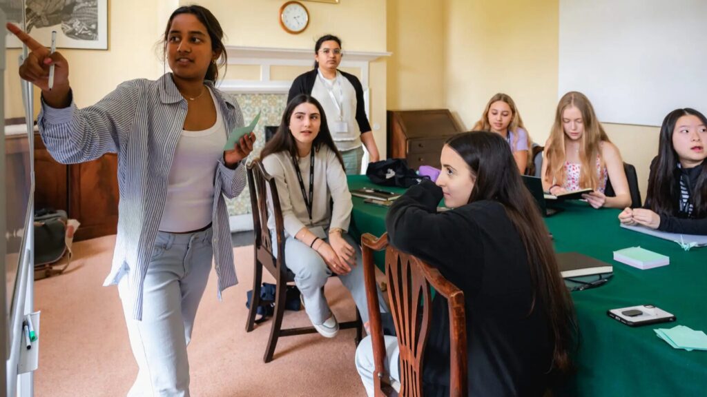 A group of high school students sitting at a rectangular table; an instructor is standing and pointing towards a whiteboard, addressing the group.