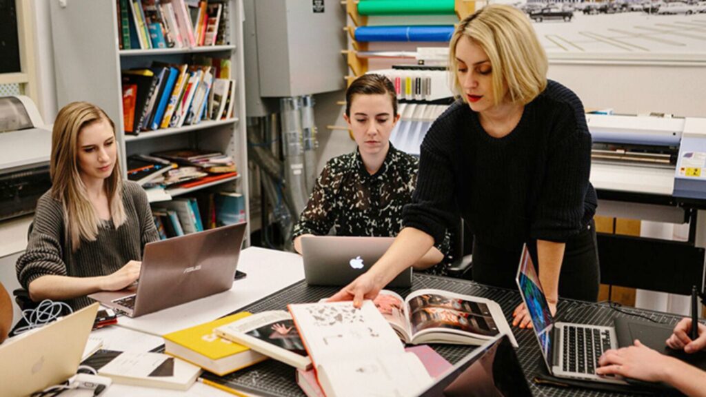 A design instructor showing a book to a table full of seated students with laptops.