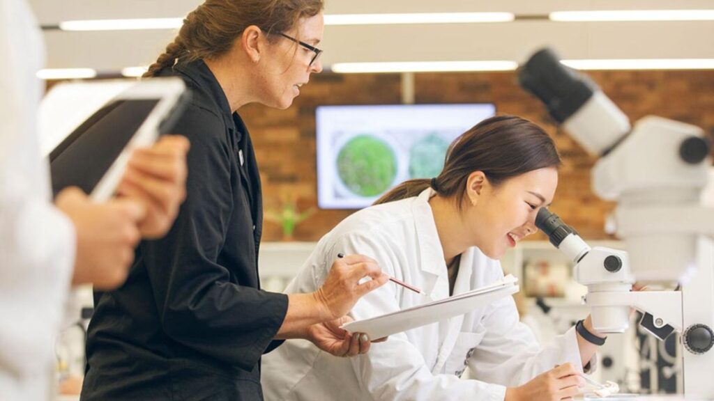 A student wearing a lab coat and peering into a microscope while an instructor stands behind and guides