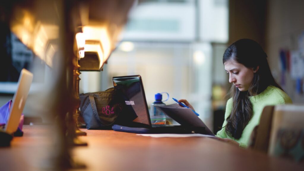 A student attending an online on their laptop and taking notes using a blue pen