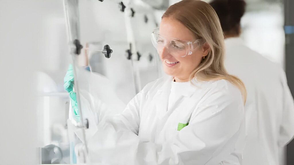 A student wearing a lab coat and green gloves and performing an experiment