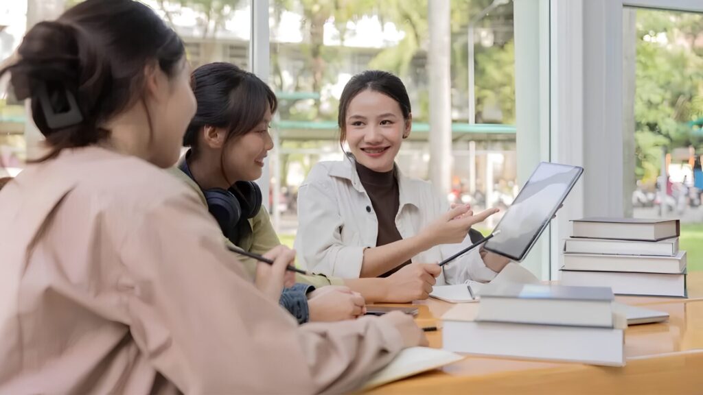 A trio of students sitting around a table lined with books and discussing something.